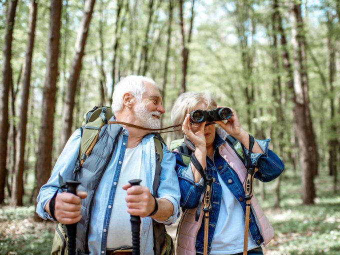 Zwei ältere Menschen laufen durch den Wald und beobachten mit dem Fernglas die Natur. 