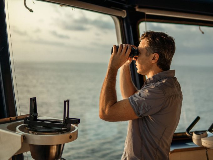 Ein Mann steht auf einem Schiff und schaut mit einem Marine-Fernglas in die Ferne.