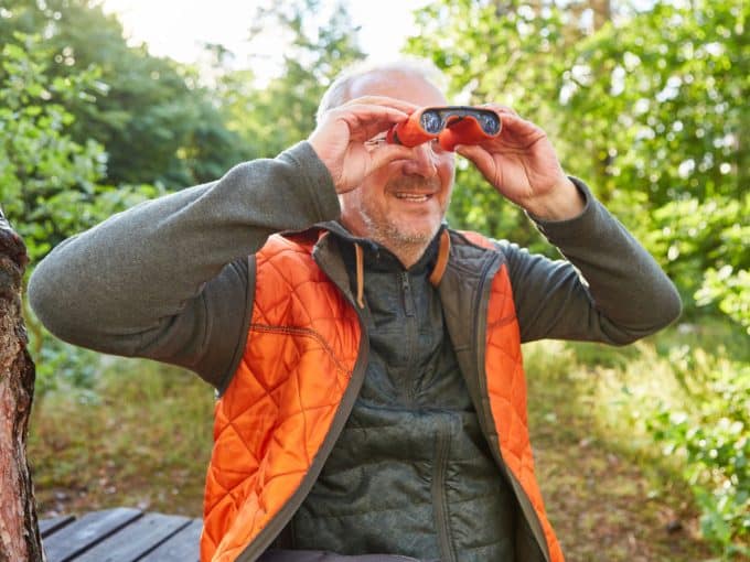 Ein Mann sitzt in einem Wald und beobachtet mit einem Fernglas etwas.