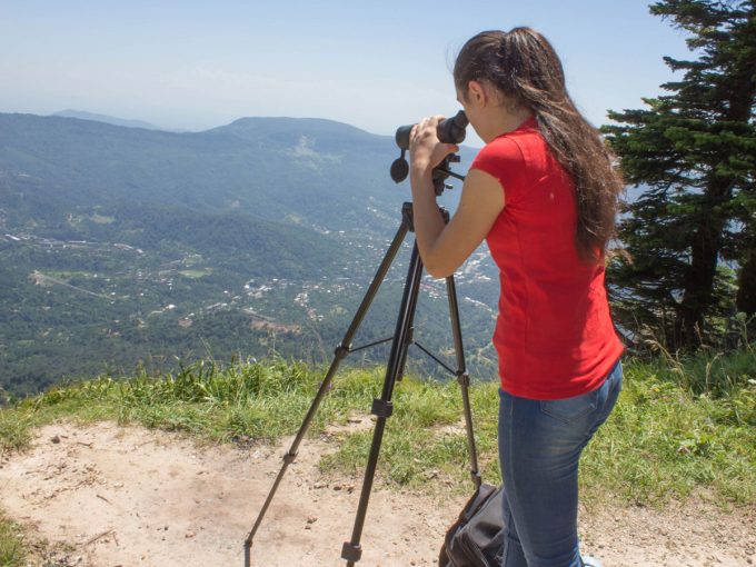 Eine Frau steht auf einem Berg und schaut durch ihr Spektiv auf ein Tal. 