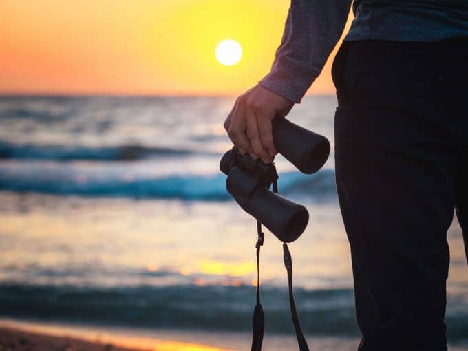 Eine Person steht am Meer und beobachtet den Sonnenuntergang mit einem Fernglas in der Hand.