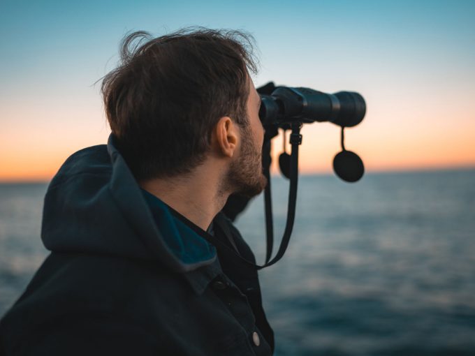 Ein Mann schaut mit einem Fernglas von der Küste aufs Meer.